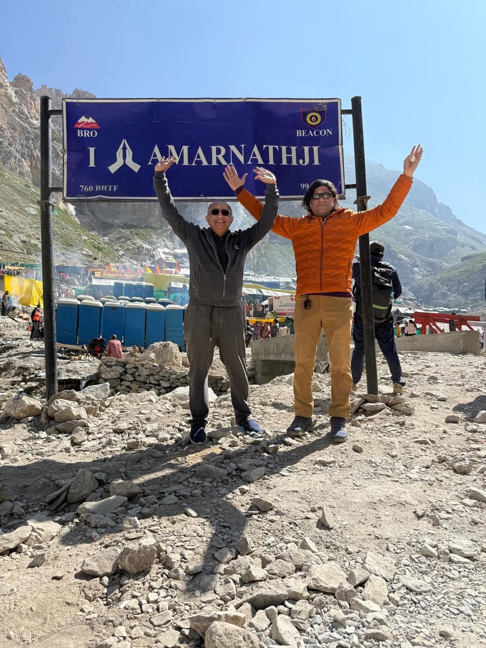 Two travellers with raised arms beneath the Amarnathji sign in the mountains